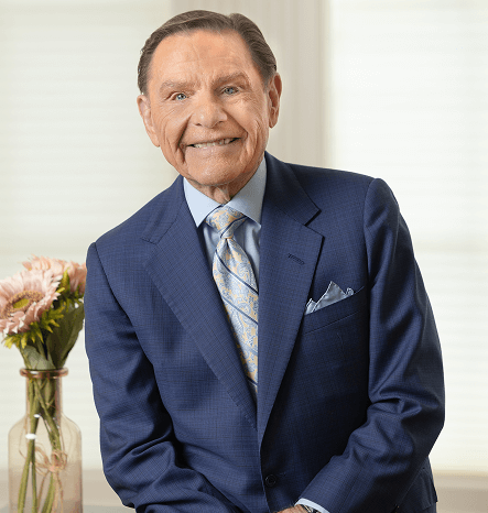 Headshot of Kenneth Copeland standing and smiling in front of a clear vase of pink roses